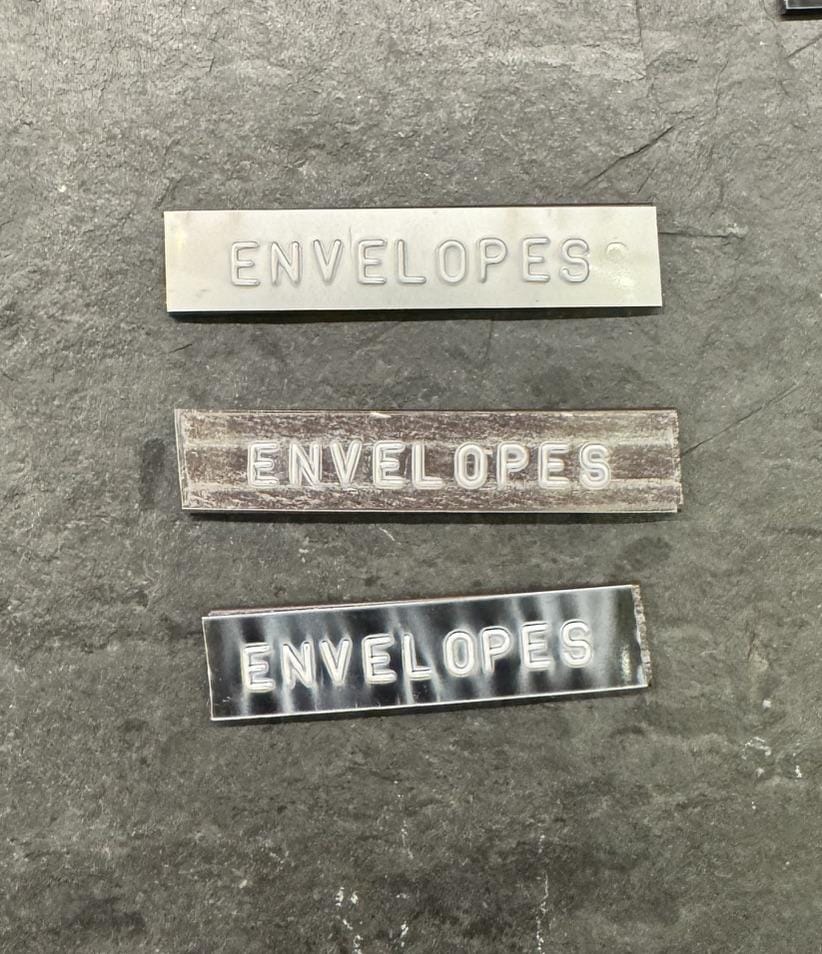 Three embossed labels reading ENVELOPES on metal, clear and black tape, laid on dark slate